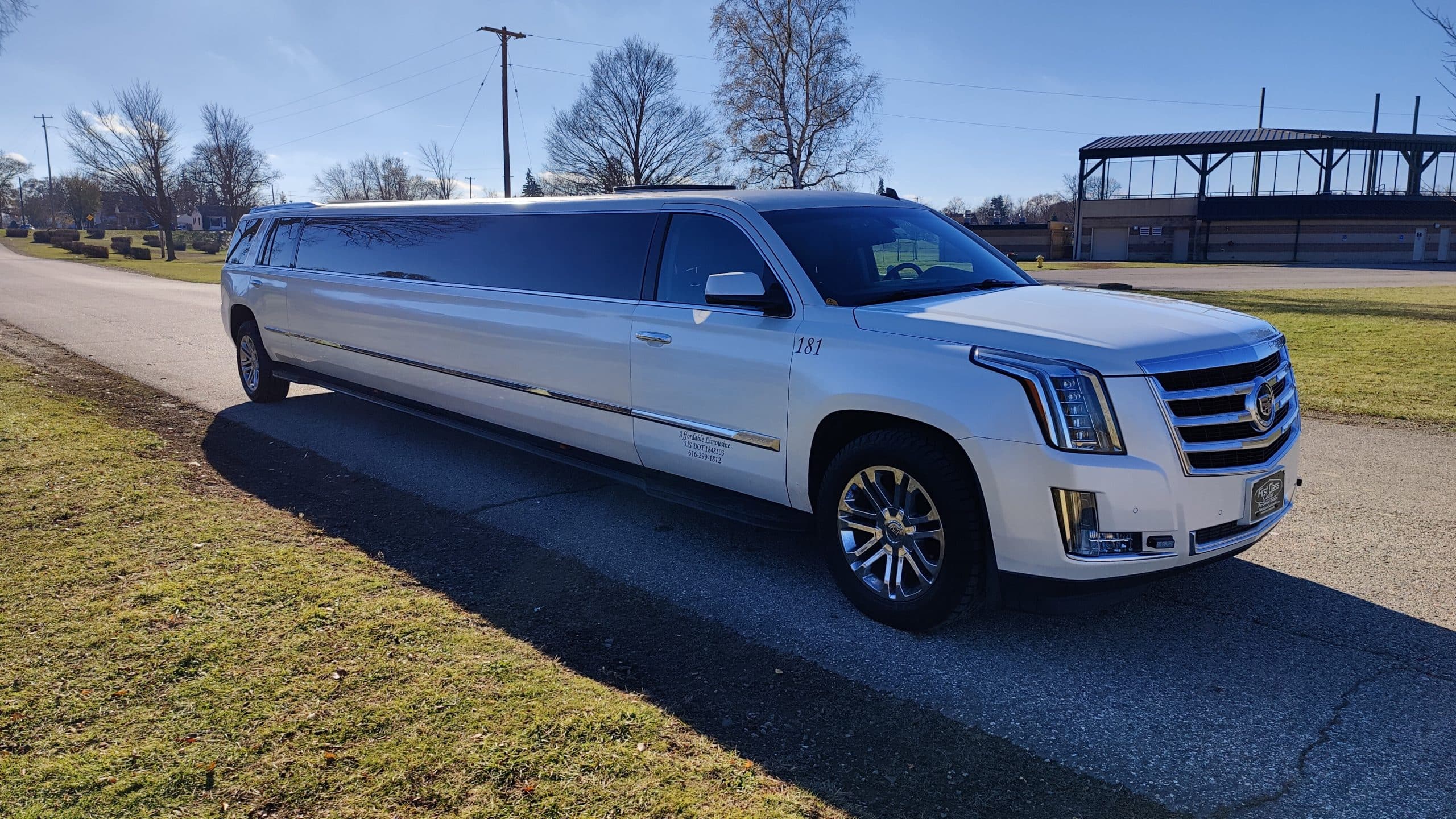 A long white stretch SUV limousine from a premier Limo Service Jacksonville is parked on a paved road next to a grassy area, with trees, utility poles, and a covered structure in the background on a sunny FL day.