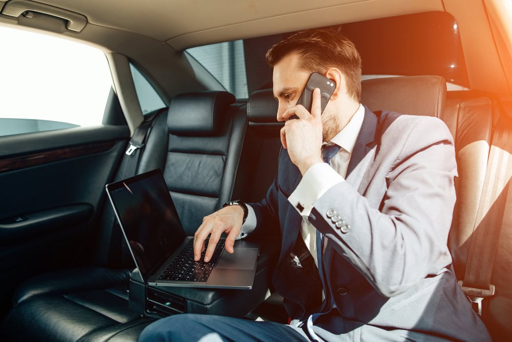 A man in a suit sits in the backseat of a FL limo service Jacksonville car, talking on his phone while working on a laptop placed on his lap as sunlight streams through the window, illuminating the interior.