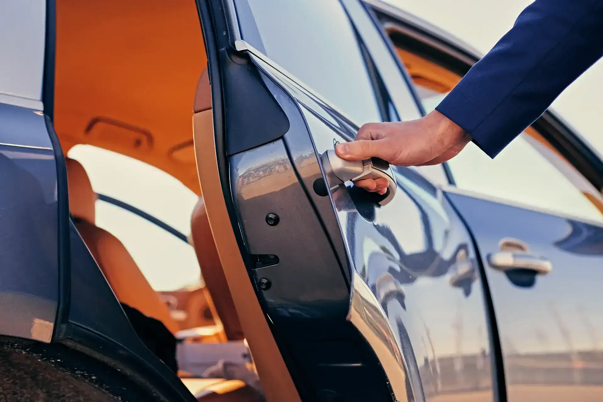A person wearing a blue suit opens the rear passenger door of a car, revealing a tan interior and sunlight reflecting off the vehicle's exterior.
